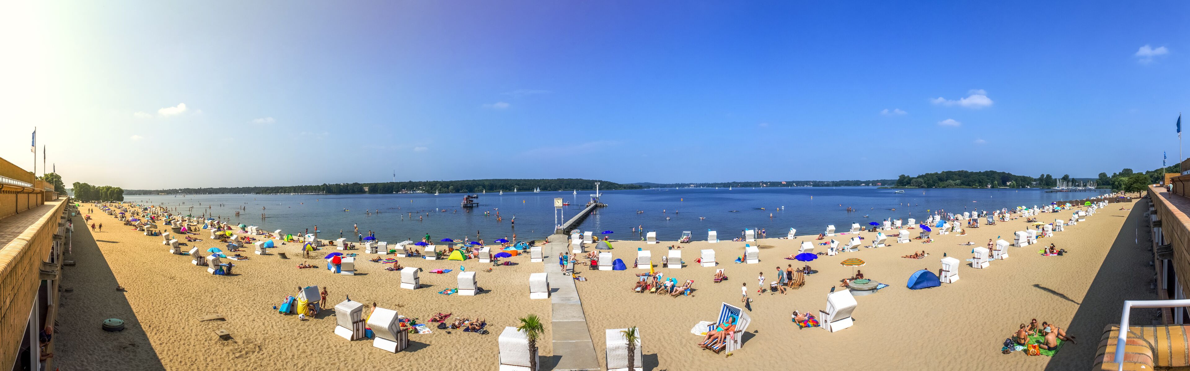 Uma praia com cadeiras de praia brancas e guarda-sóis, um píer que se estende para a água, e um céu azul claro.