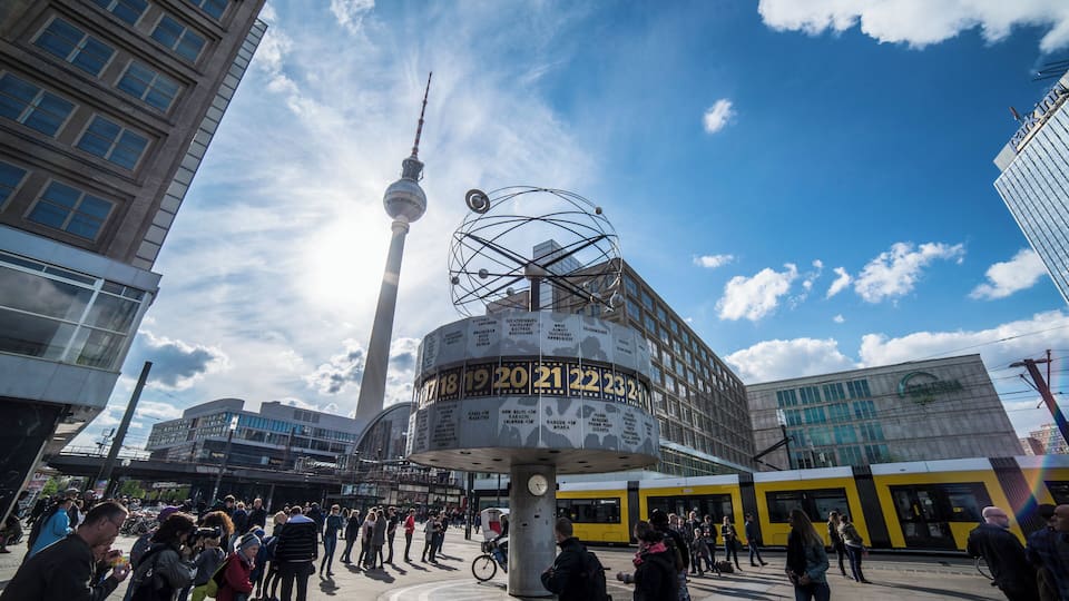 A busy urban square with a prominent tower, a circular structure with numbers, and a yellow tram.