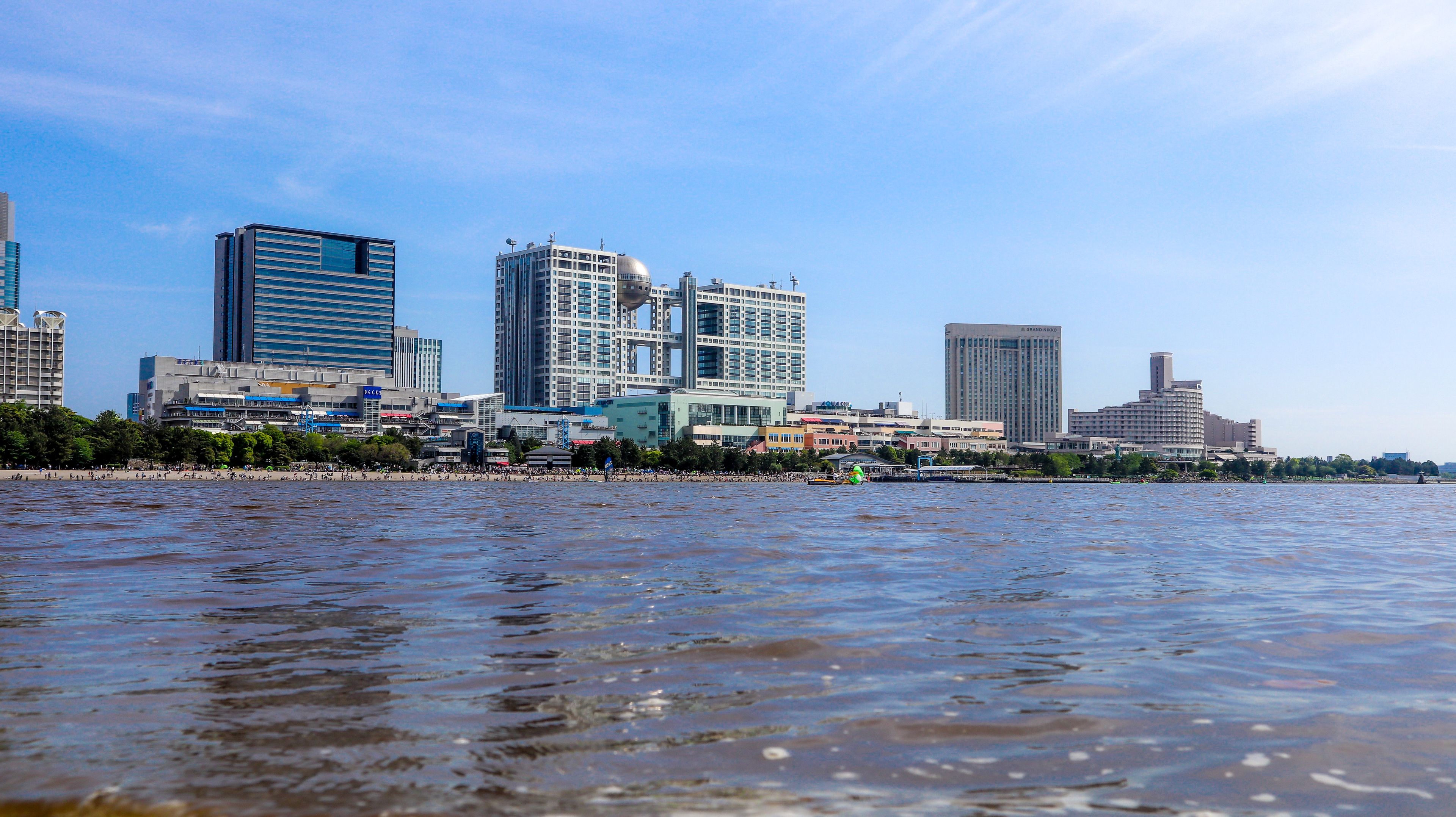 A waterfront cityscape with modern buildings, a beach, and calm water.