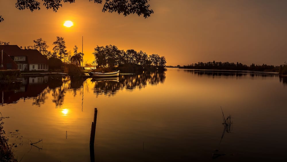 Une rivière paisible au coucher du soleil, avec une maison et un bateau sur la rive gauche.