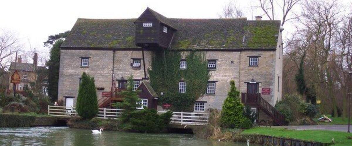 A historic stone building with a weir, surrounded by a grassy area and a river.
