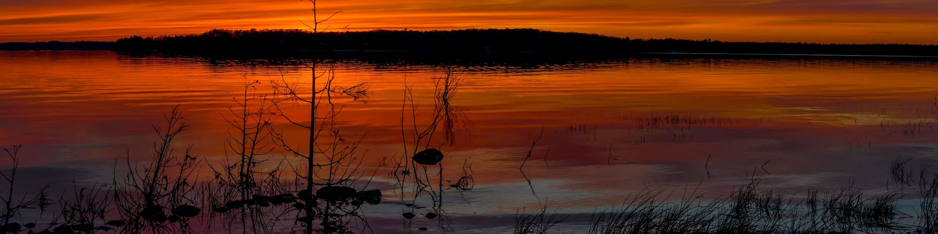 A sunset over a calm lake with silhouetted trees and a distant shoreline.