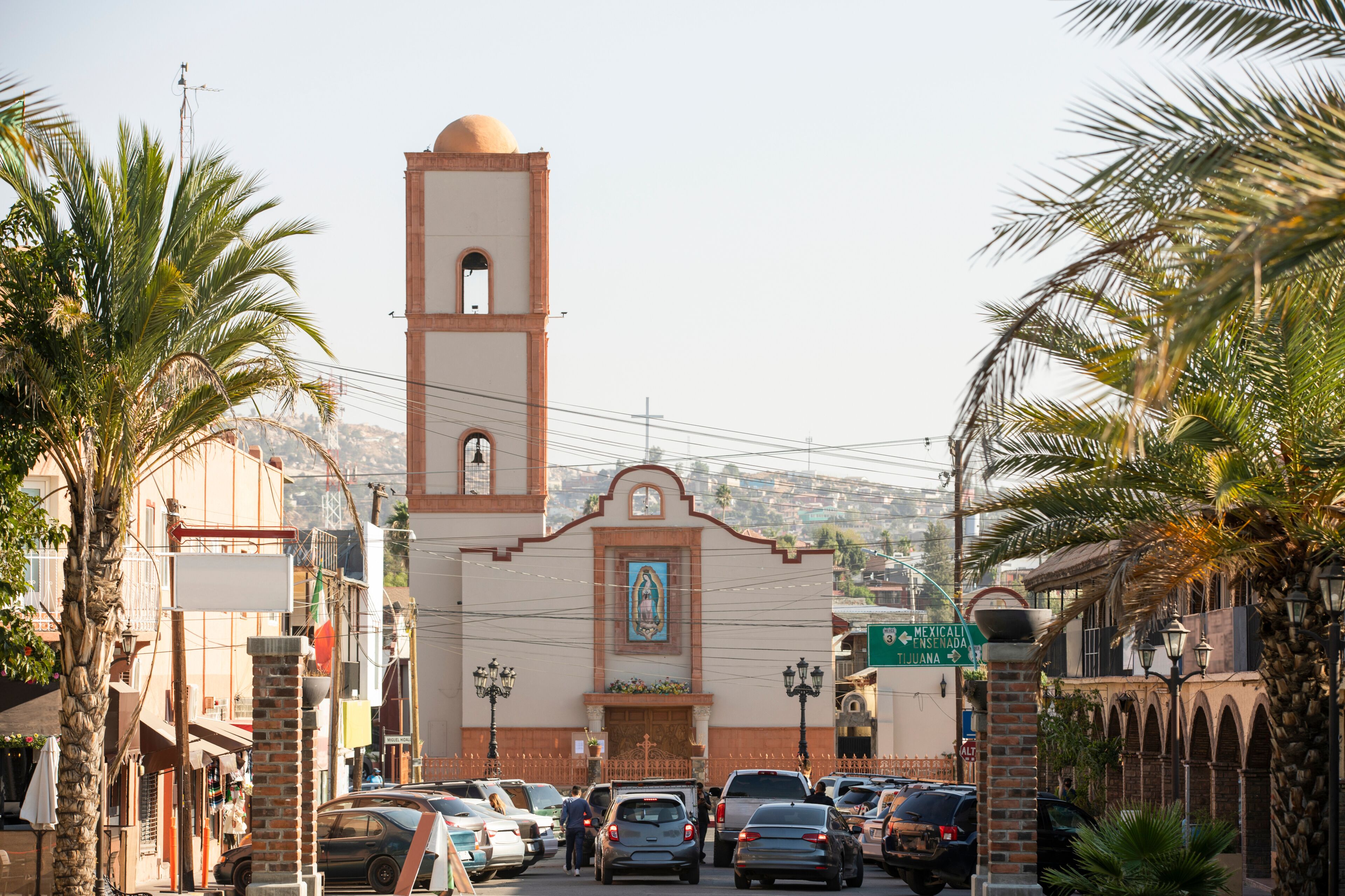 A church with a bell tower, palm trees, parked cars, and a street sign.
