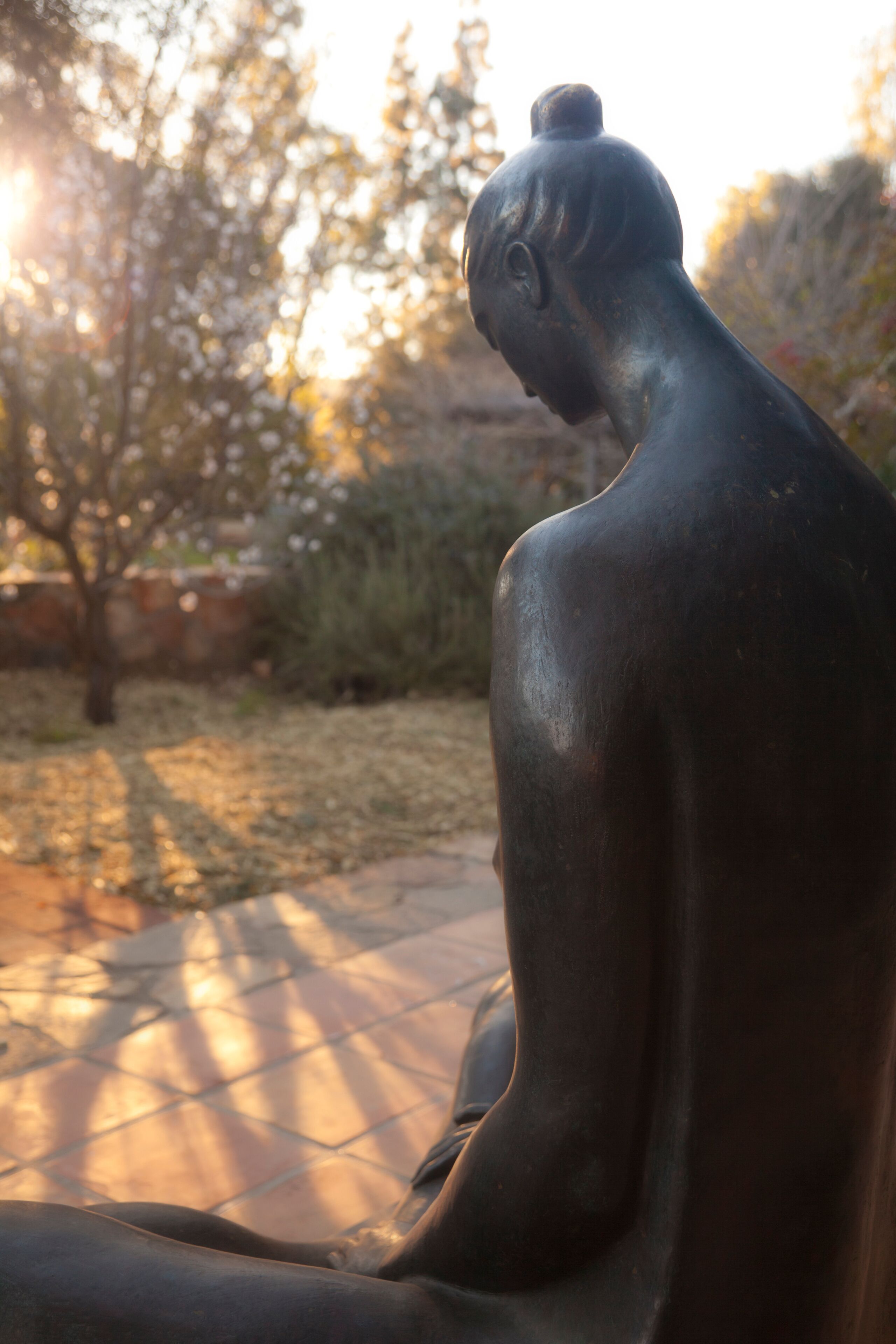 Una estatua de bronce de una figura sentada en un jardín con árboles y suelo de baldosas.