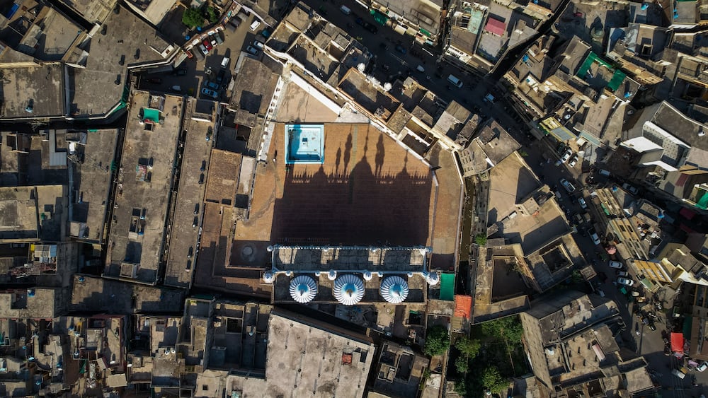 Une vue aĂ©rienne dâune zone urbaine densĂ©ment peuplĂ©e, avec des rues Ă©troites, une grande place ouverte et un bĂątiment imposant surmontĂ© dâun toit bleu.