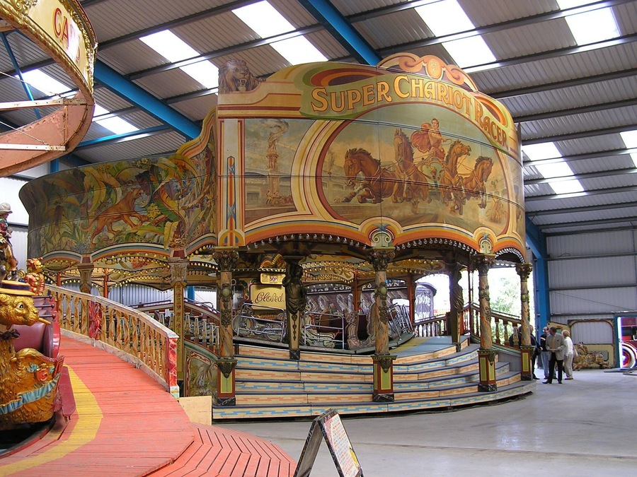 Edwards’ Super Chariot Racer at Dingles Fairground Heritage Centre, Lifton, Devon Robert Edwards ordered the ride new in 1934 at a cost of £1,750. It was built in Burton on Trent by Messrs. Orton, Sons and Spooner. The ride is 42 feet in diameter, plus the porch and steps, and was decorated in art deco ‘jazz’ style, popular in the 1930s. After the war the ride was rebuilt with the massive Chariot Racer scene on the front boards. This was constructed at Ortons and painted by their artists, Sid and Albert Howell. The Chariot Racer was travelled by the Edwards family until 1991 when it was placed in store in Swindon until acquired by the Fairground Heritage Trust. It arrived at the site in 2006 and with the help of Frank Edwards, it was assembled within a fortnight of arriving at its new home, and opened to the public later that year. fairground-heritage.org.uk/