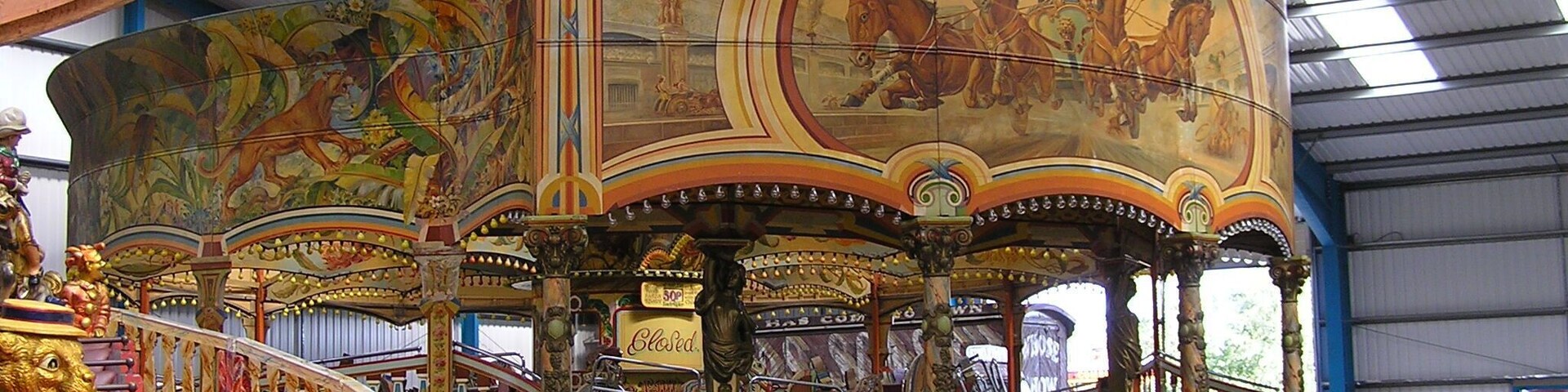 Edwards’ Super Chariot Racer at Dingles Fairground Heritage Centre, Lifton, Devon Robert Edwards ordered the ride new in 1934 at a cost of £1,750. It was built in Burton on Trent by Messrs. Orton, Sons and Spooner. The ride is 42 feet in diameter, plus the porch and steps, and was decorated in art deco ‘jazz’ style, popular in the 1930s. After the war the ride was rebuilt with the massive Chariot Racer scene on the front boards. This was constructed at Ortons and painted by their artists, Sid and Albert Howell. The Chariot Racer was travelled by the Edwards family until 1991 when it was placed in store in Swindon until acquired by the Fairground Heritage Trust. It arrived at the site in 2006 and with the help of Frank Edwards, it was assembled within a fortnight of arriving at its new home, and opened to the public later that year. fairground-heritage.org.uk/