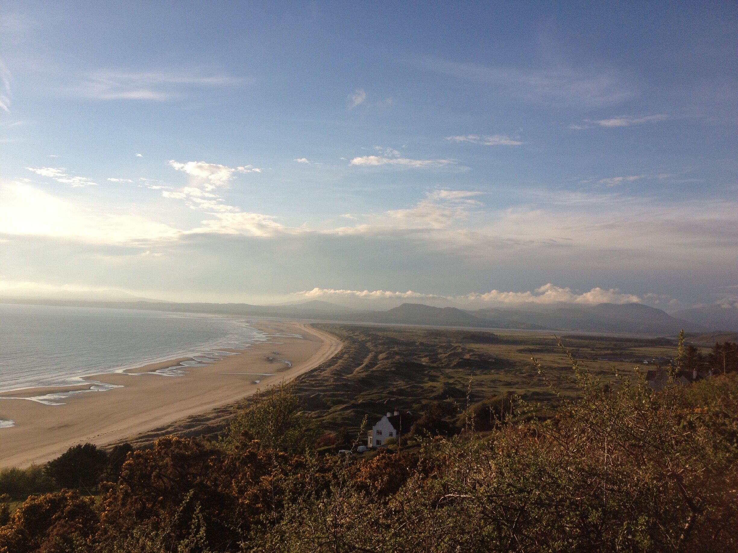 A coastal landscape with a sandy beach, rolling hills, and a white house in the foreground.