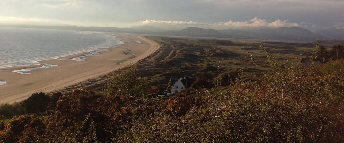 A coastal landscape with a sandy beach, rolling hills, and a white house in the foreground.