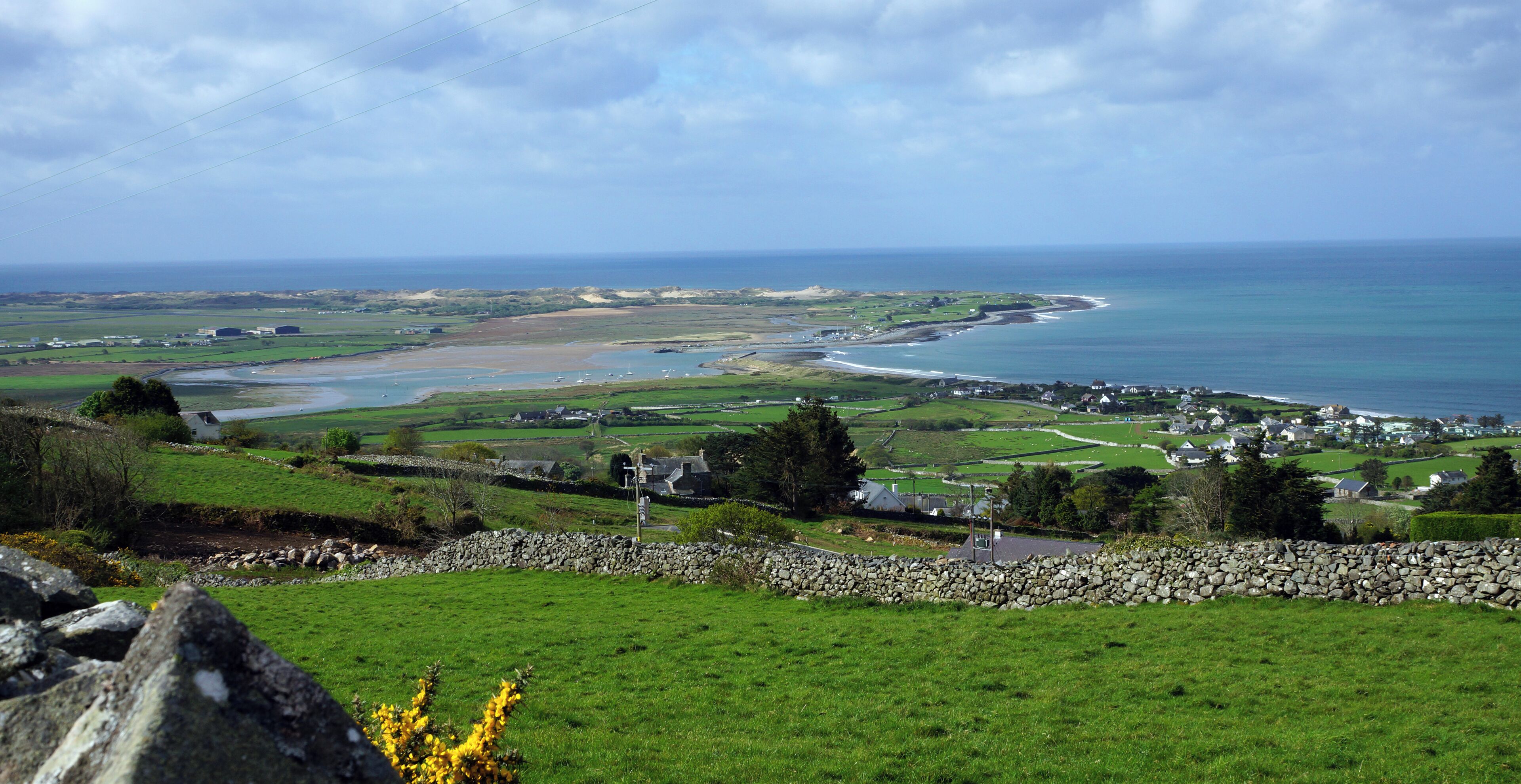 A coastal landscape with green fields, a stone wall, and a view of the sea and distant buildings.