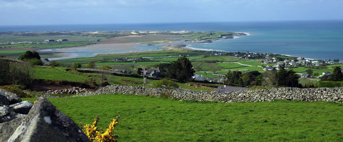 A coastal landscape with green fields, a stone wall, and a view of the sea and distant buildings.
