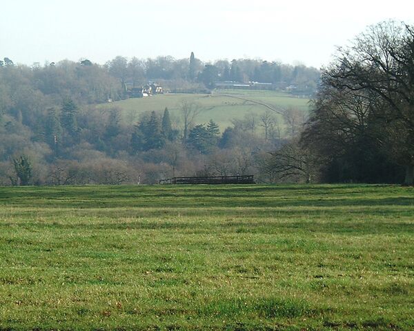 A rural landscape with a field, a wooden structure, and a hill with trees and buildings in the distance.