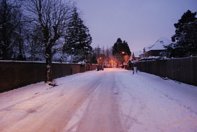 A snow-covered road with houses on both sides, a brick wall, and bare trees.