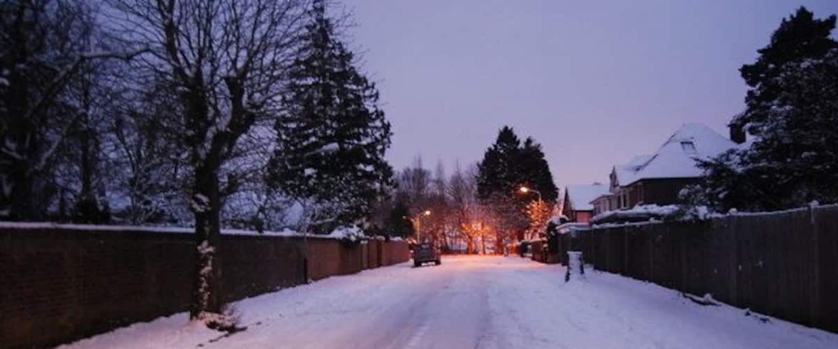 A snow-covered road with houses on both sides, a brick wall, and bare trees.