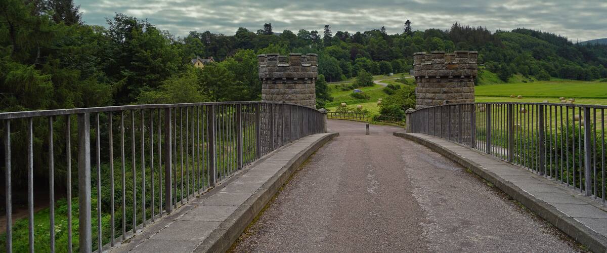 A stone bridge with metal railings, surrounded by lush greenery and a cloudy sky.