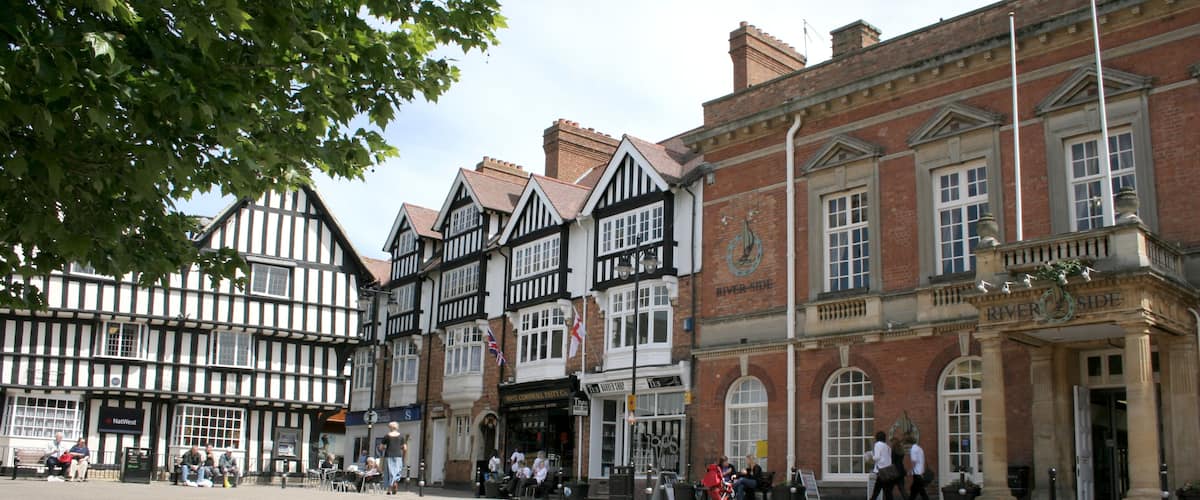 A historic town square with Tudor-style buildings, a River Side sign, and people walking.
