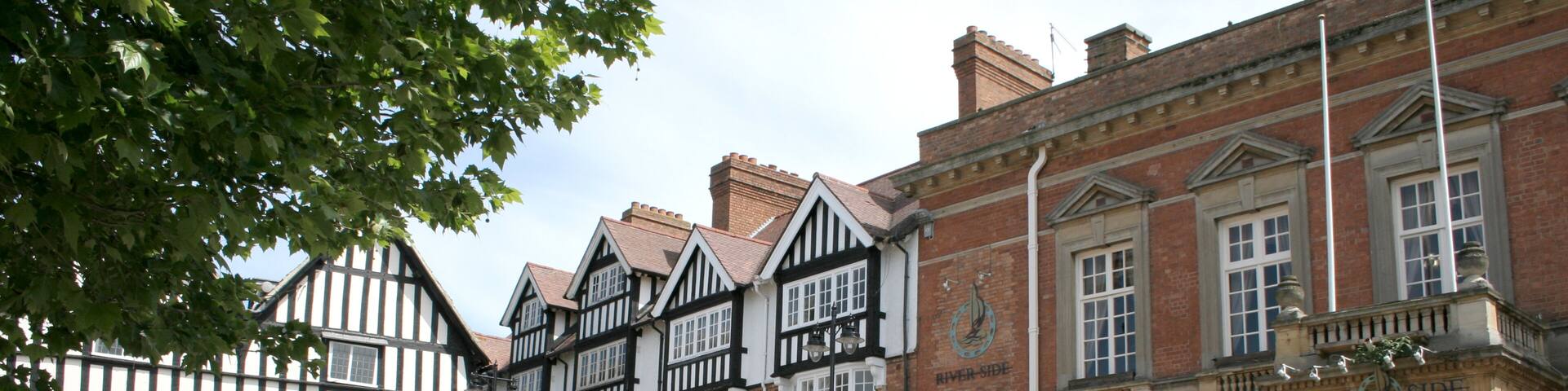 A historic town square with Tudor-style buildings, a River Side sign, and people walking.