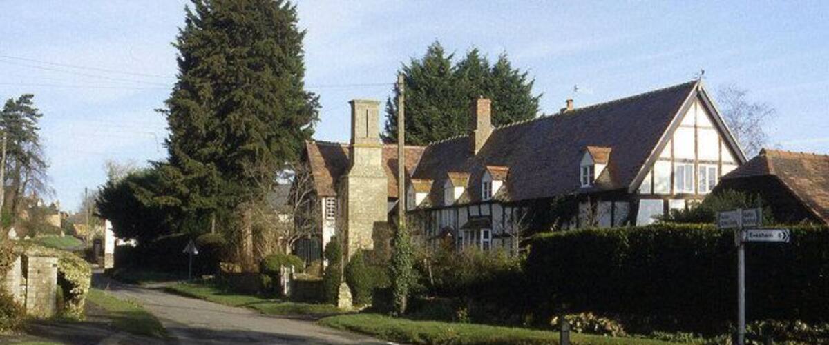A quiet village street with traditional half-timbered houses, a large tree, and a clear blue sky.