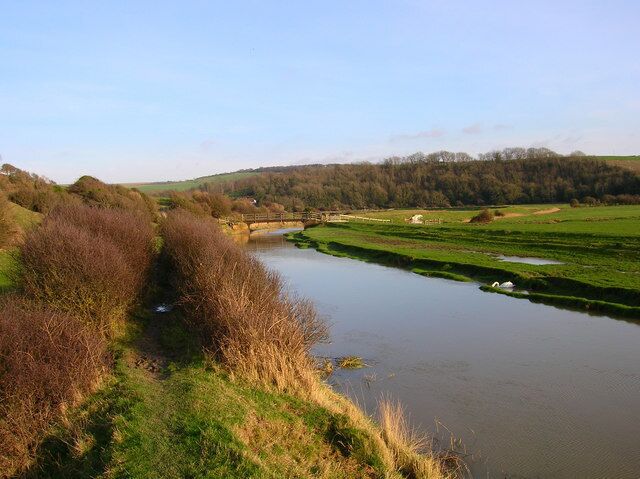 A river flowing through a rural landscape with grassy banks and a forested hill in the background.