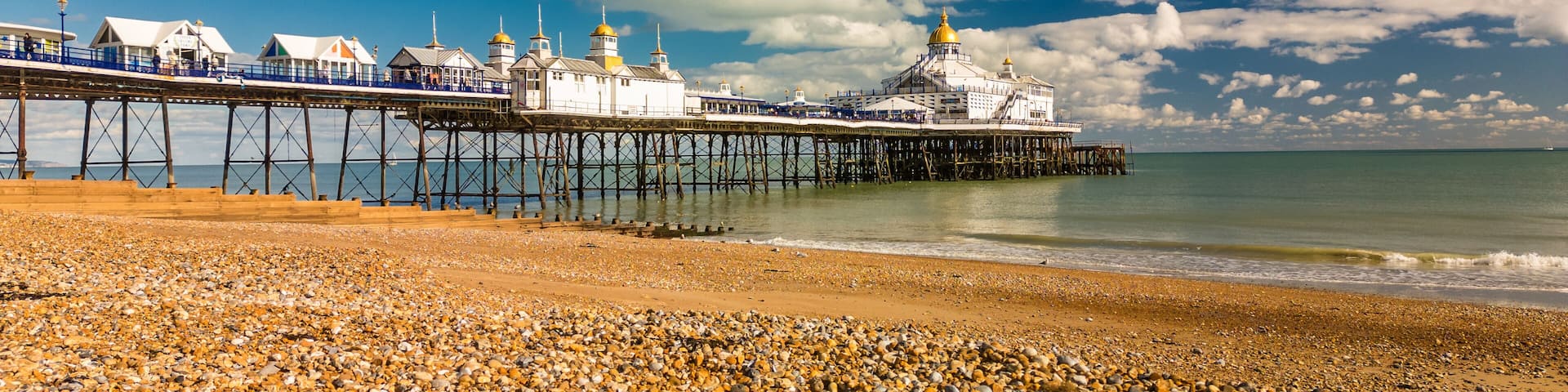 A pebbled beach with a pier extending into the sea, featuring a large building with golden domes.