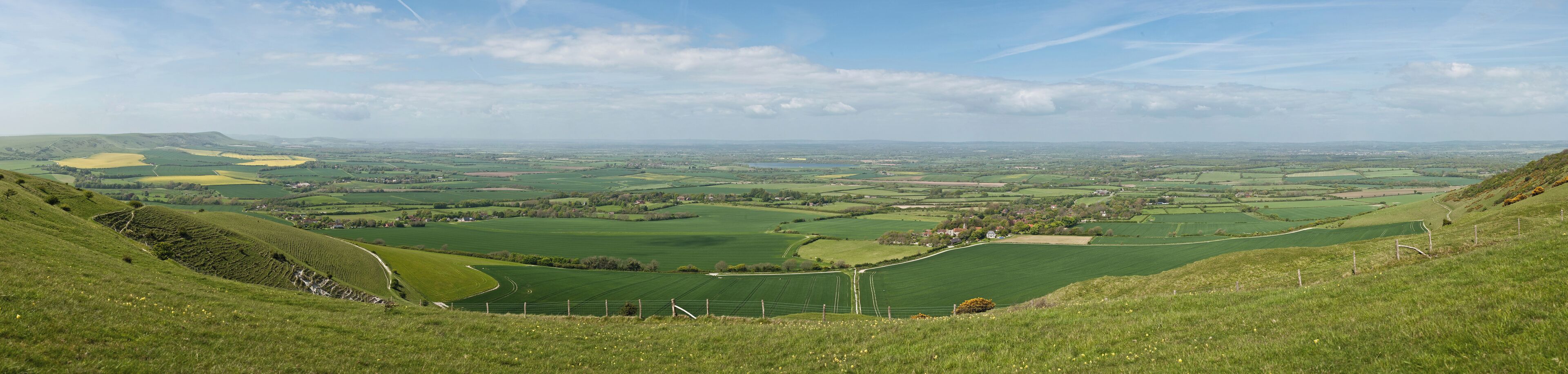 A panoramic wide view of the Long Man civil parish in East Sussex, England as viewed from just above the Long Man of Wilmington (at the bottom of the hill not visible). On the far left is the village of Milton Street, in the centre-right is the village of Wilmington, and in the distance on the far-right is Hailsham.