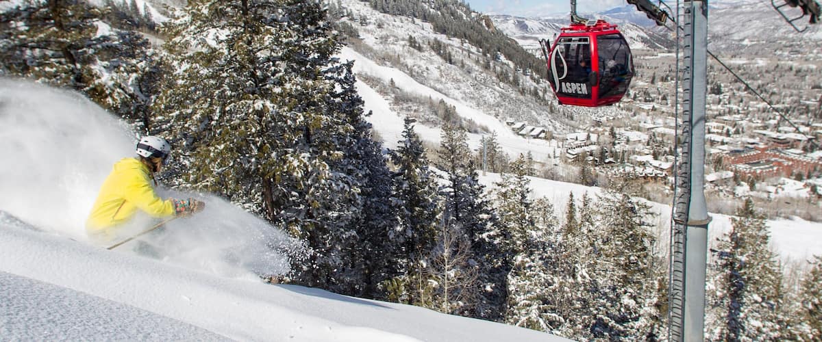 A ski lift with a red cabin labeled "ASPEN" in a snowy mountainous area with trees and a clear sky.