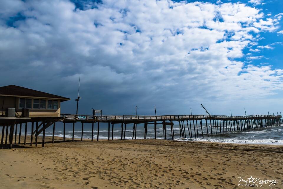 A beach with a wooden pier extending into the ocean, a lifeguard tower, and a cloudy sky.