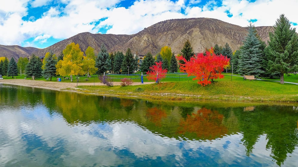 A calm lake with reflections, a wooden bridge, autumn-colored trees, and mountains in the background.