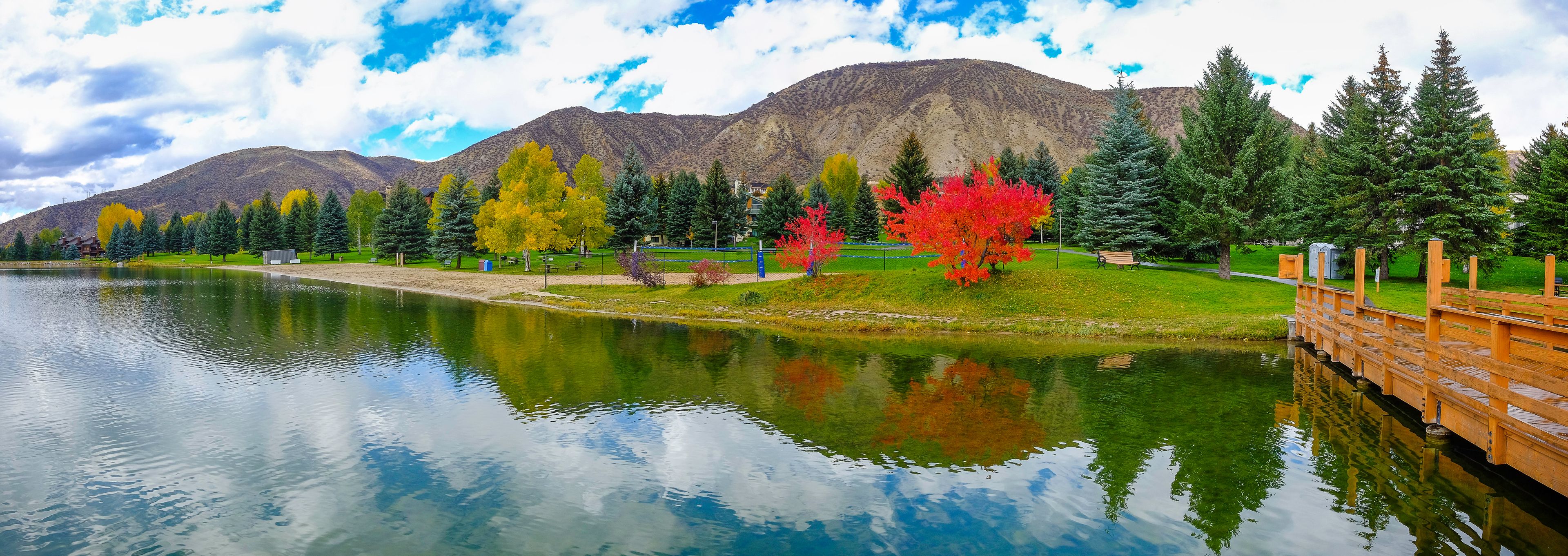 A calm lake with reflections, a wooden bridge, autumn-colored trees, and mountains in the background.