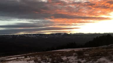 Um pôr do sol sobre uma paisagem montanhosa com picos nevados e um vale florestado.