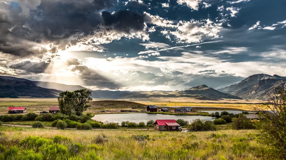 A rural landscape with a lake, red-roofed cabins, and mountains under a cloudy sky.
