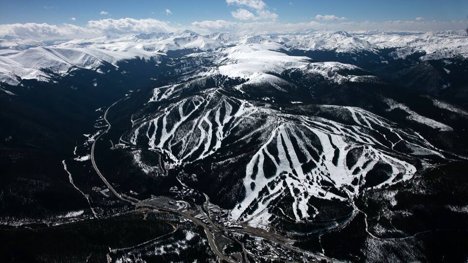 A snow-covered mountain range with distinct ridges and valleys, a winding road, and a clear blue sky.