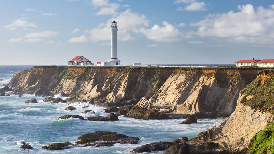 A coastal lighthouse, rocky cliffs, and a row of houses along the cliff edge.