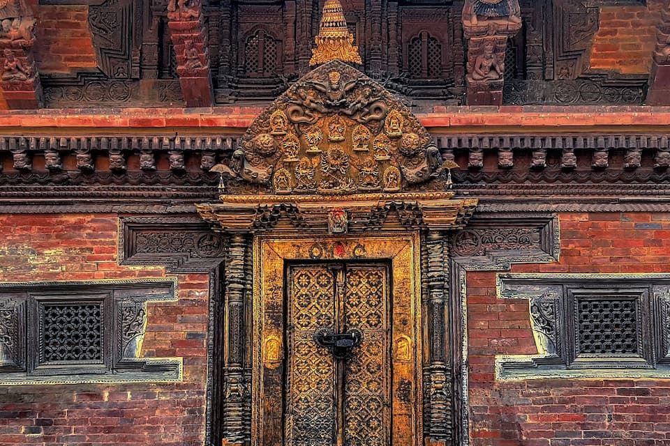 An intricately carved wooden door with a golden frame, set against a brick wall with detailed carvings and a small hanging light bulb.