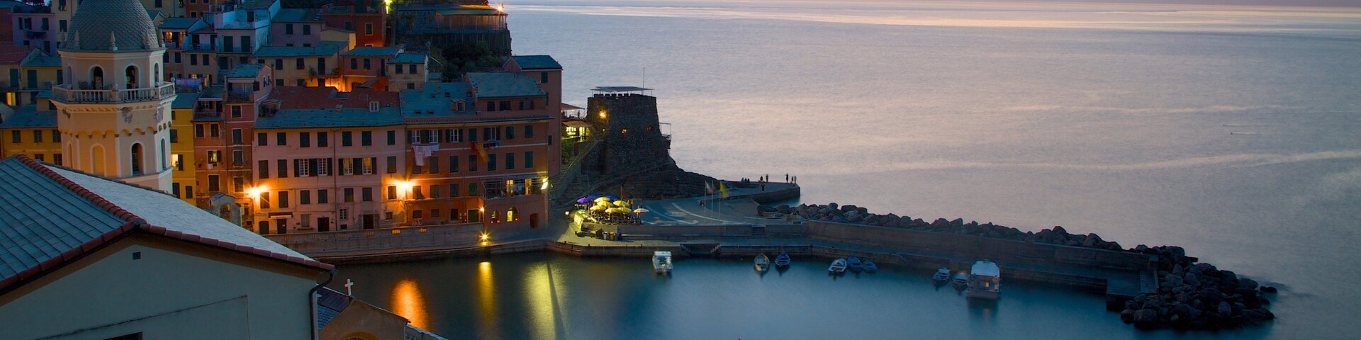 A coastal town at sunset with historic buildings, a fortified tower, and calm waters.