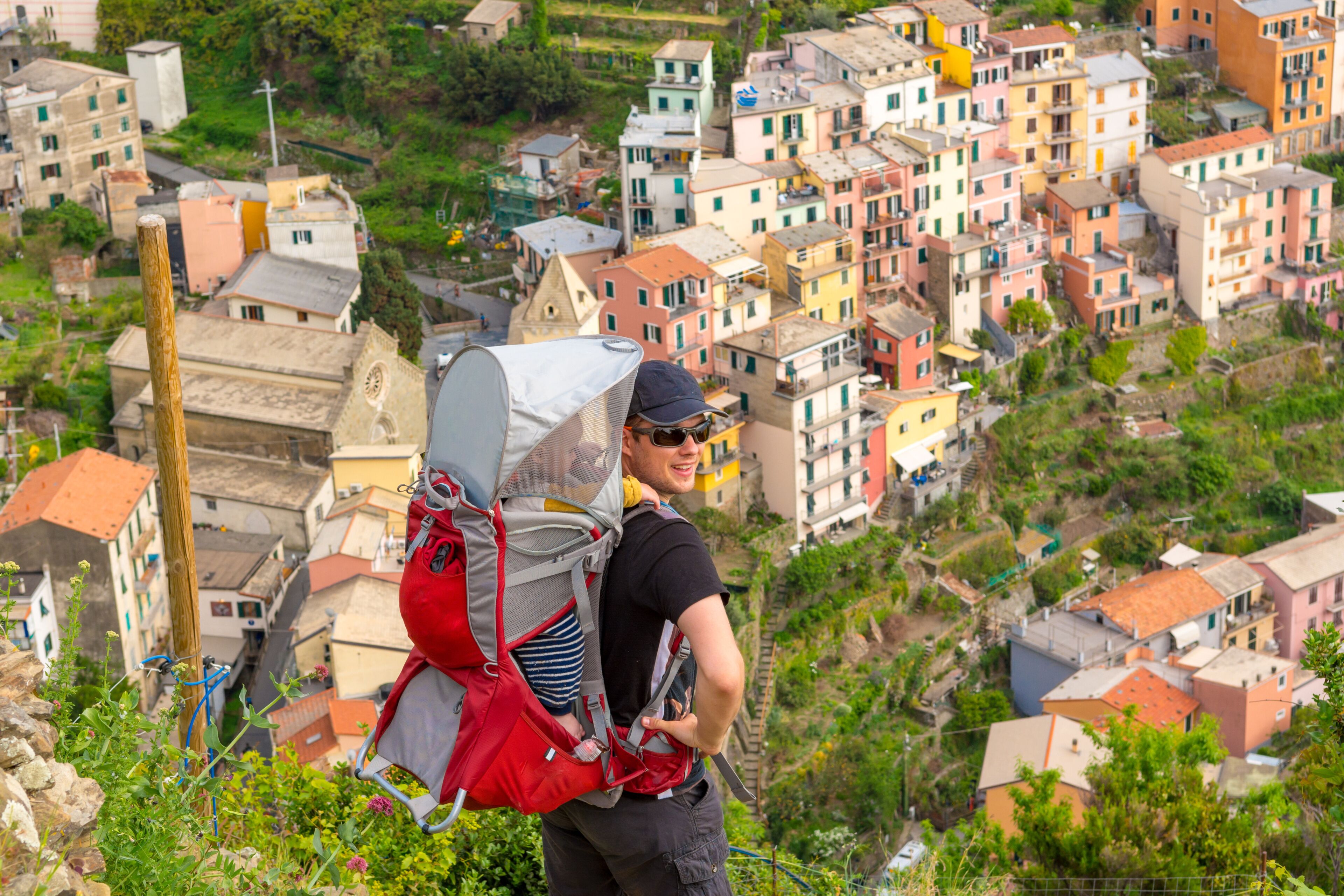 Una persona con una mochila grande lleva a un niño, observando un pueblo con casas apretadas y coloridas en una ladera.
