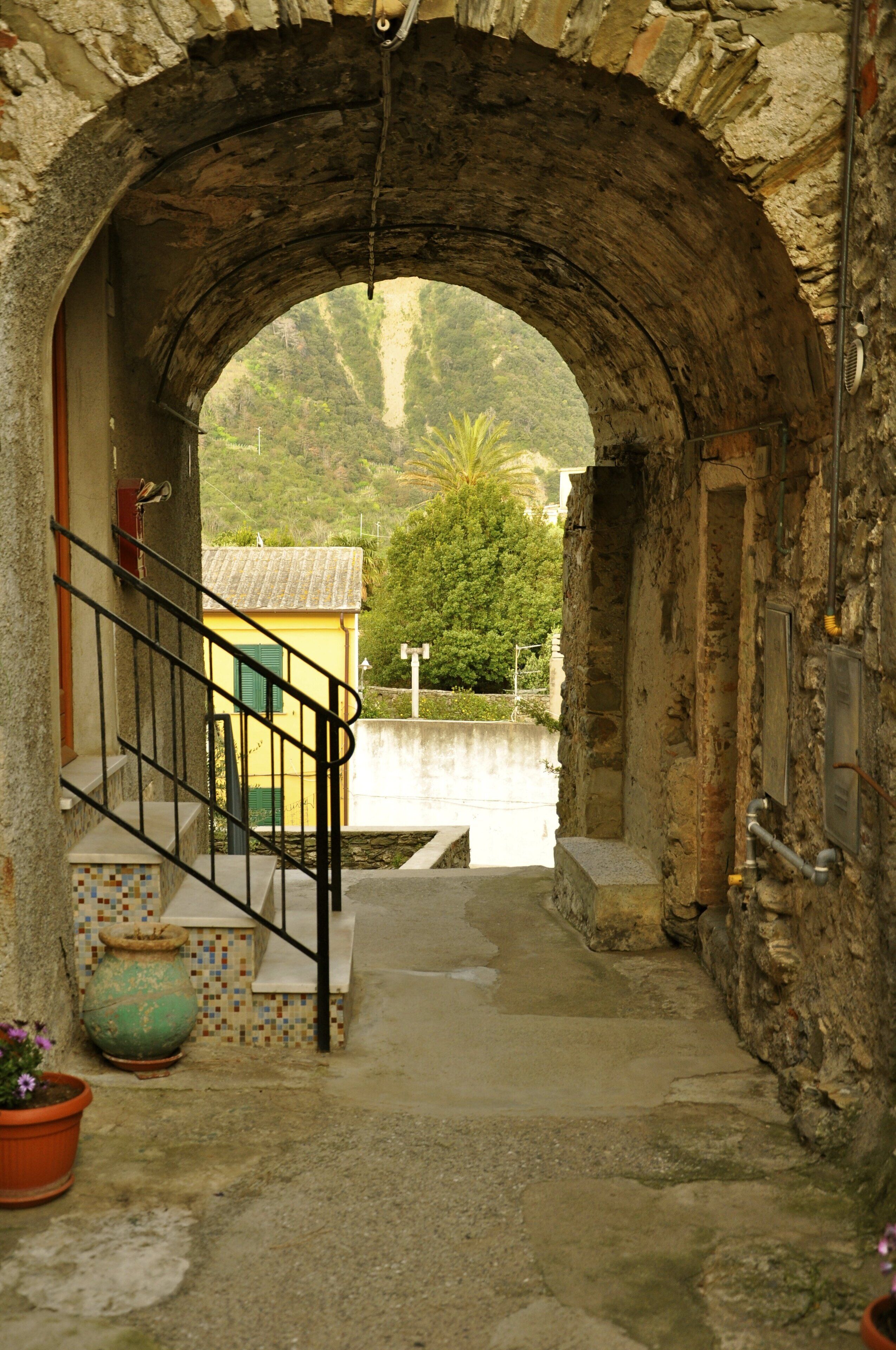 Un arco de piedra que da paso a una pequeña casa amarilla con suelo de baldosas y una puerta verde.