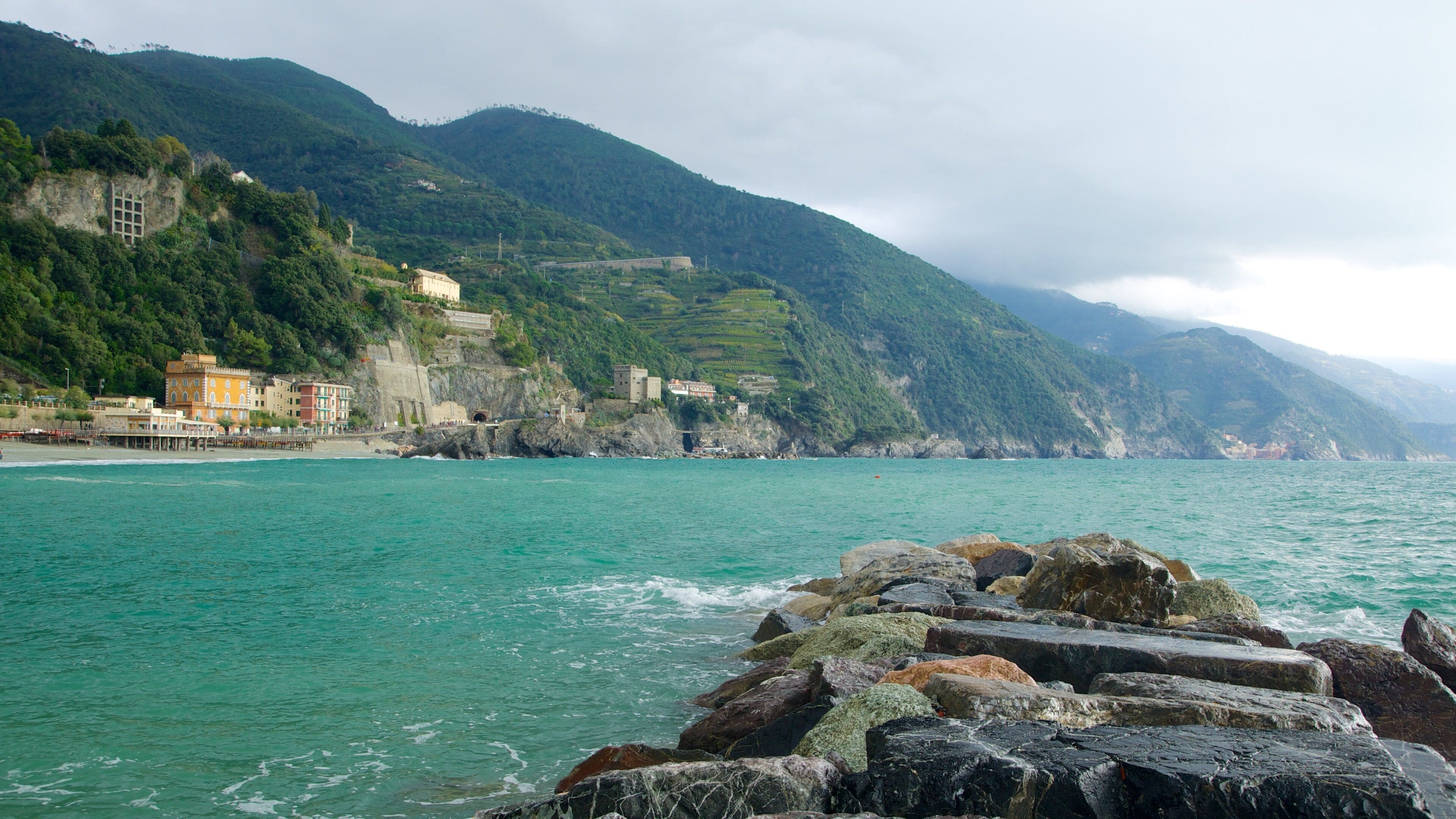A coastal town with buildings on a hillside, rocky shoreline, and turquoise sea.