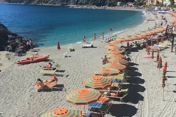 Une plage bordée de rangées de parasols rayés et de chaises longues, avec une eau d’un bleu limpide et un paysage montagneux en toile de fond.
