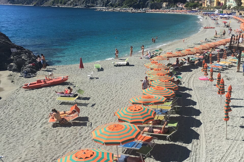 A beach with rows of striped umbrellas and sun loungers, clear blue water, and a mountainous backdrop.