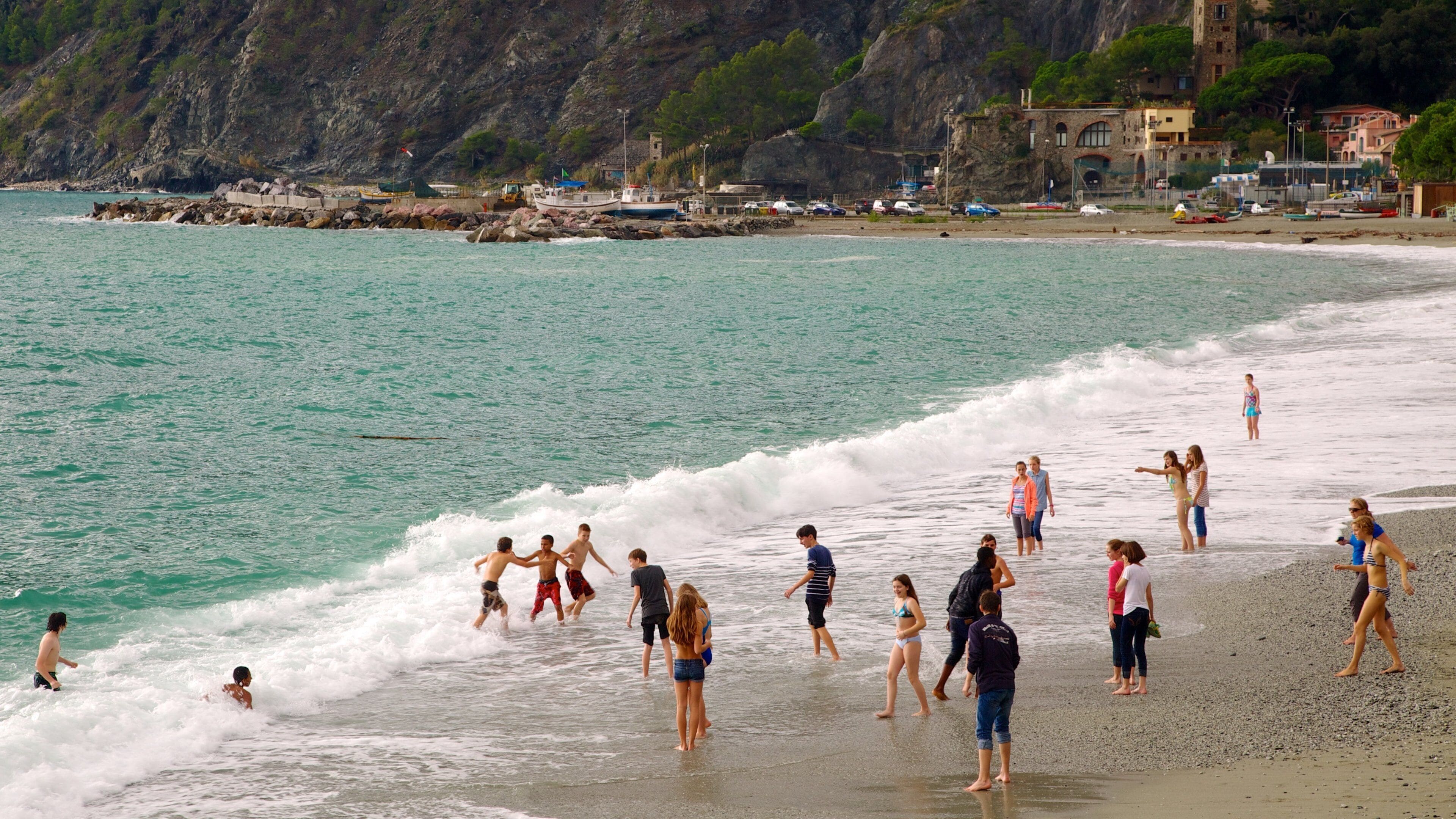 A beach with people swimming and walking along the shore, rocky cliffs in the background, and buildings near the water.