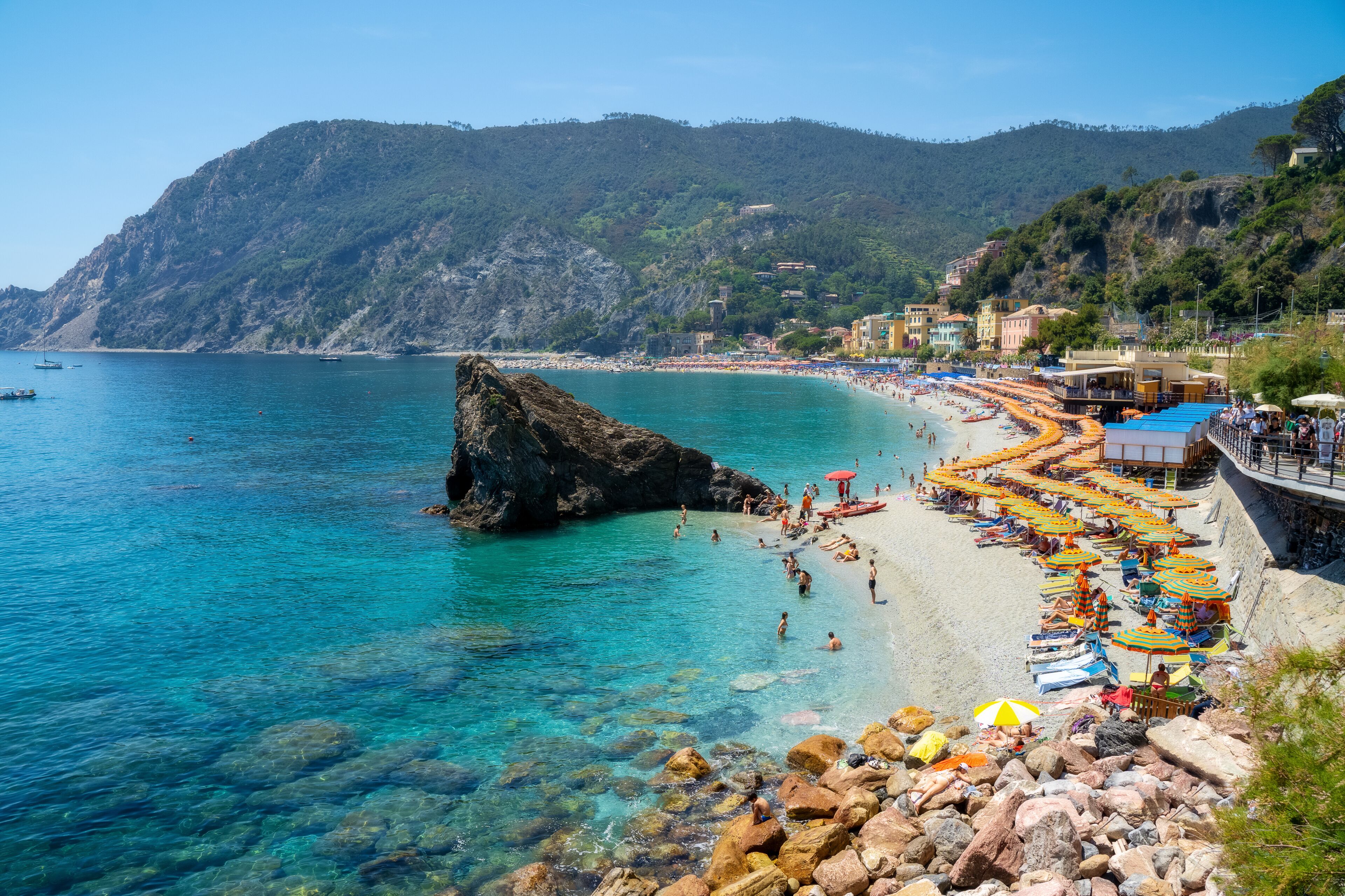 A coastal beach with clear turquoise waters, rocky shore, and a mountainous backdrop.