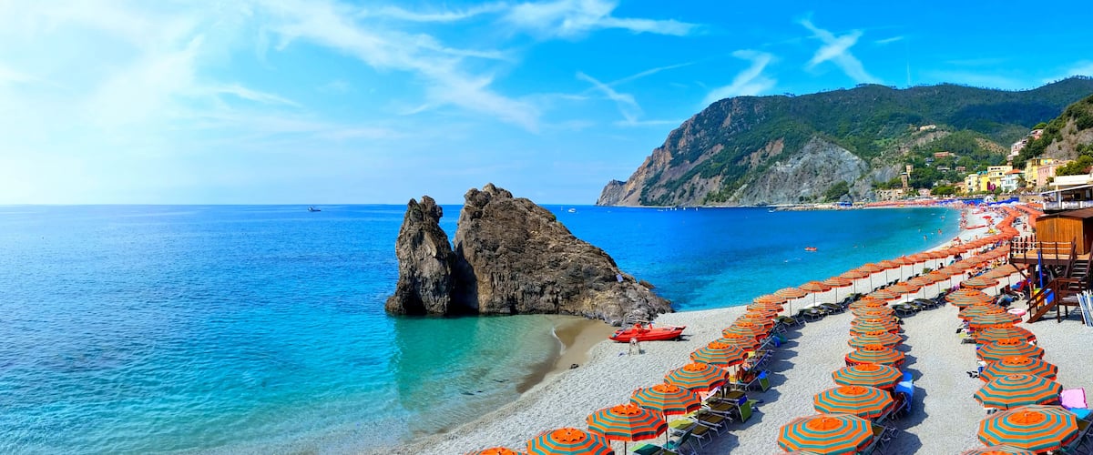 A beach with clear blue waters, rocky formations, and rows of colorful umbrellas.