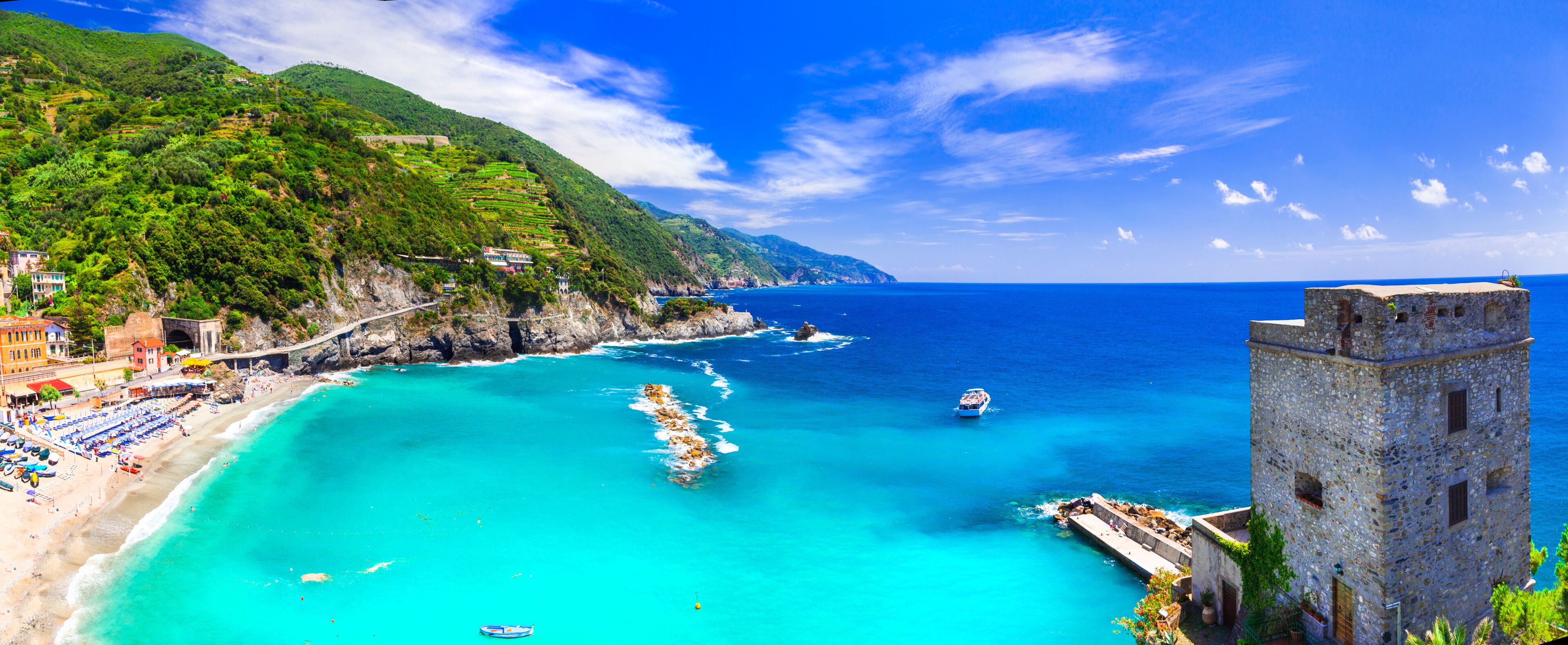 A coastal landscape with a turquoise sea, terraced vineyards, a stone tower, and a beach with umbrellas.