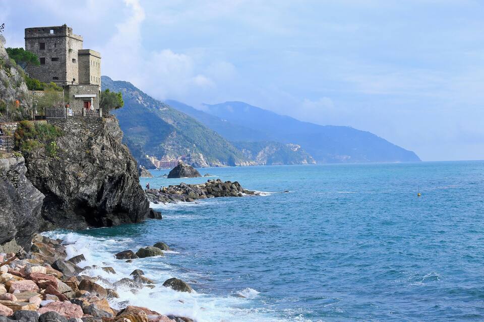 A coastal cliff with a historic stone tower, rocky shoreline, and a clear view of the sea and distant mountains.