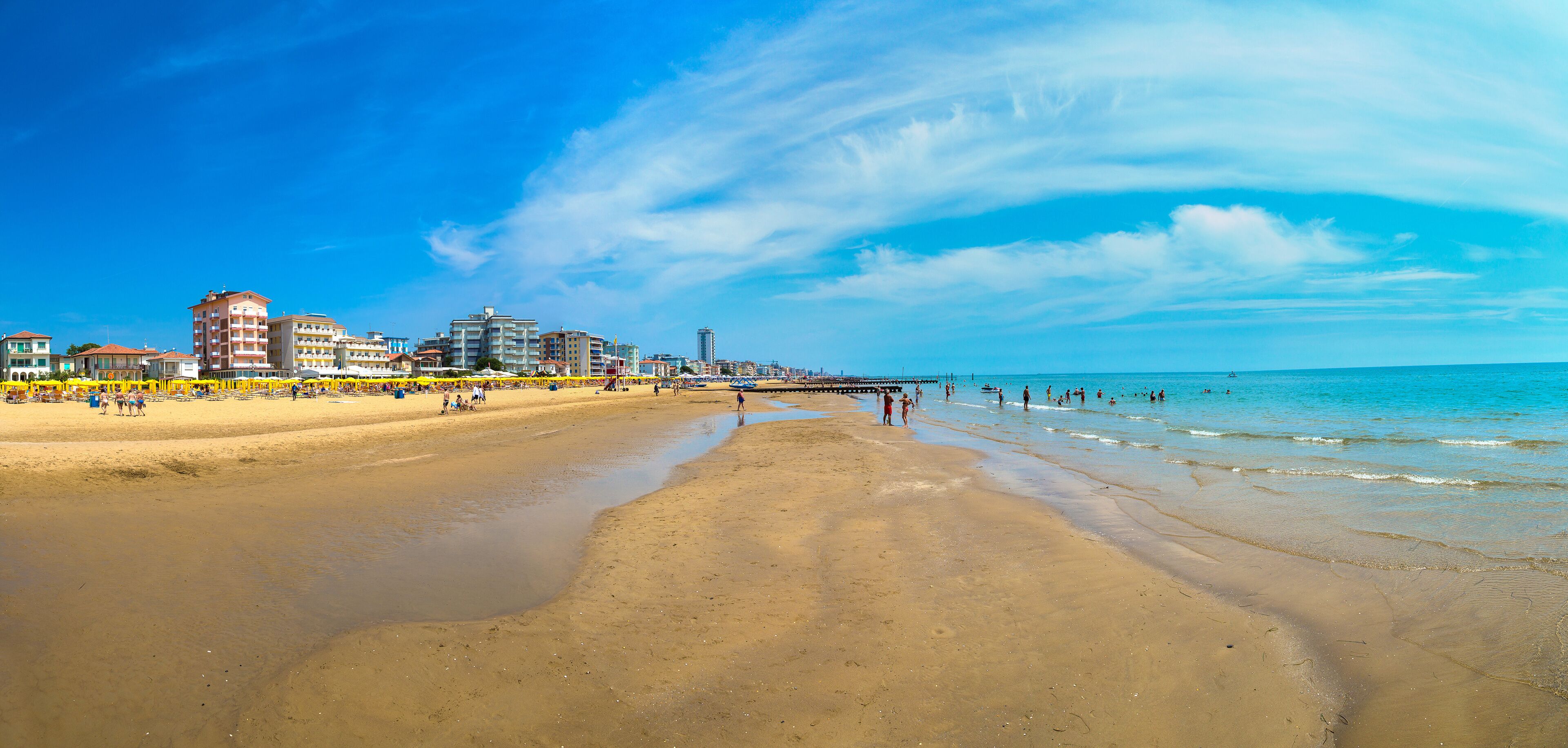 A beach with a pier extending into the water, buildings along the shore, and clear skies.