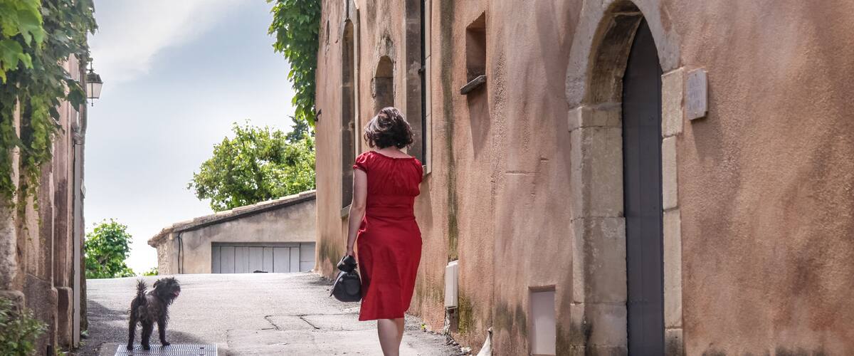 A woman in a red dress walking a dog down a narrow street flanked by old buildings with arched windows and a tree in the background.
