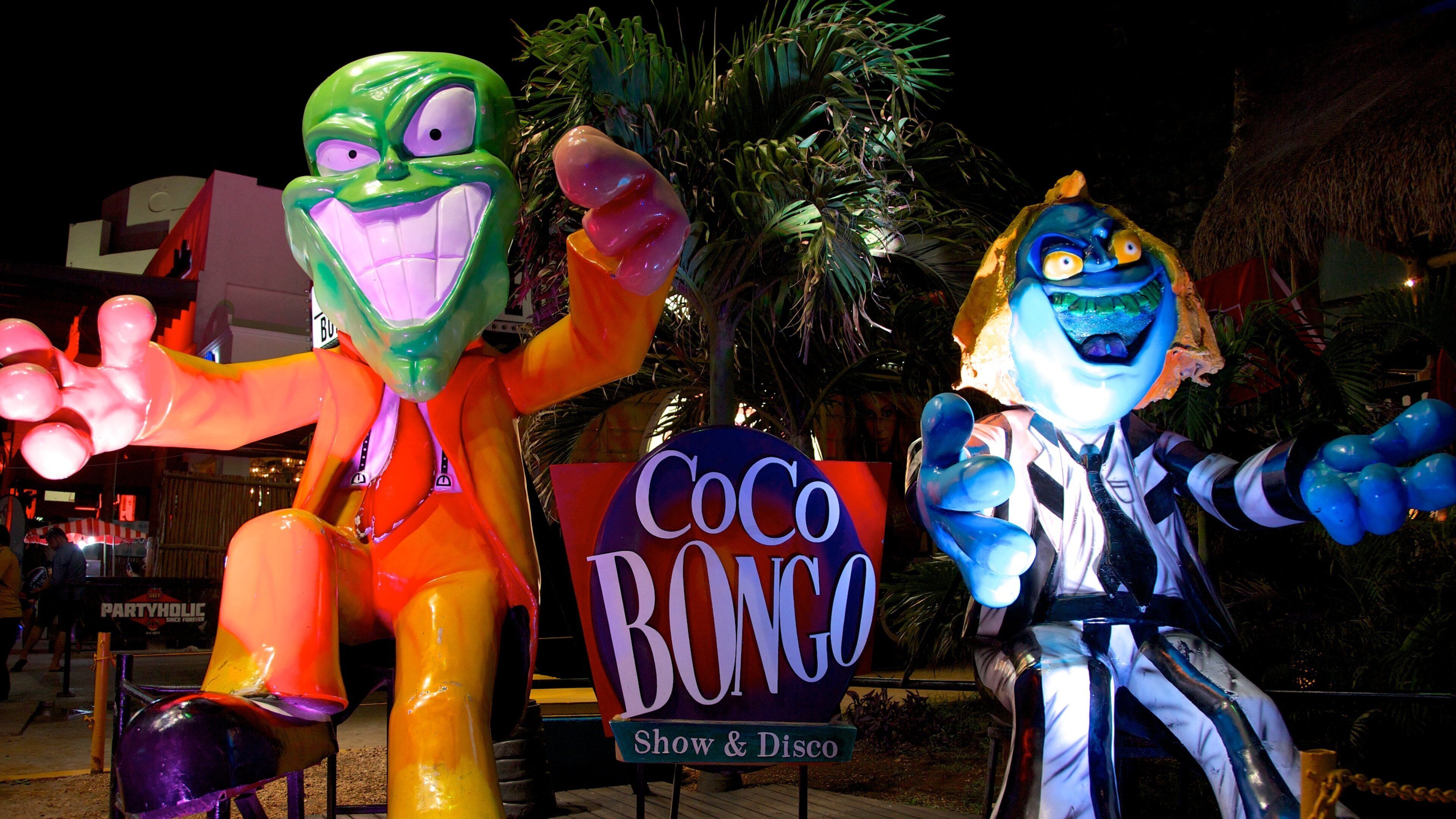Deux statues colorées représentant des personnages se trouvent devant une enseigne sur laquelle on peut lire : « Coco Bongo Show & Disco ».
