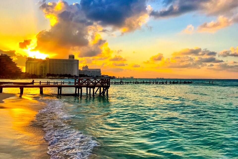 A sunset over a beach with a pier extending into the water, buildings in the background, and a clear sky.