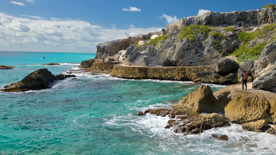 A rocky coastline with clear turquoise waters, a person walking on a stone path, and a cliff with vegetation.